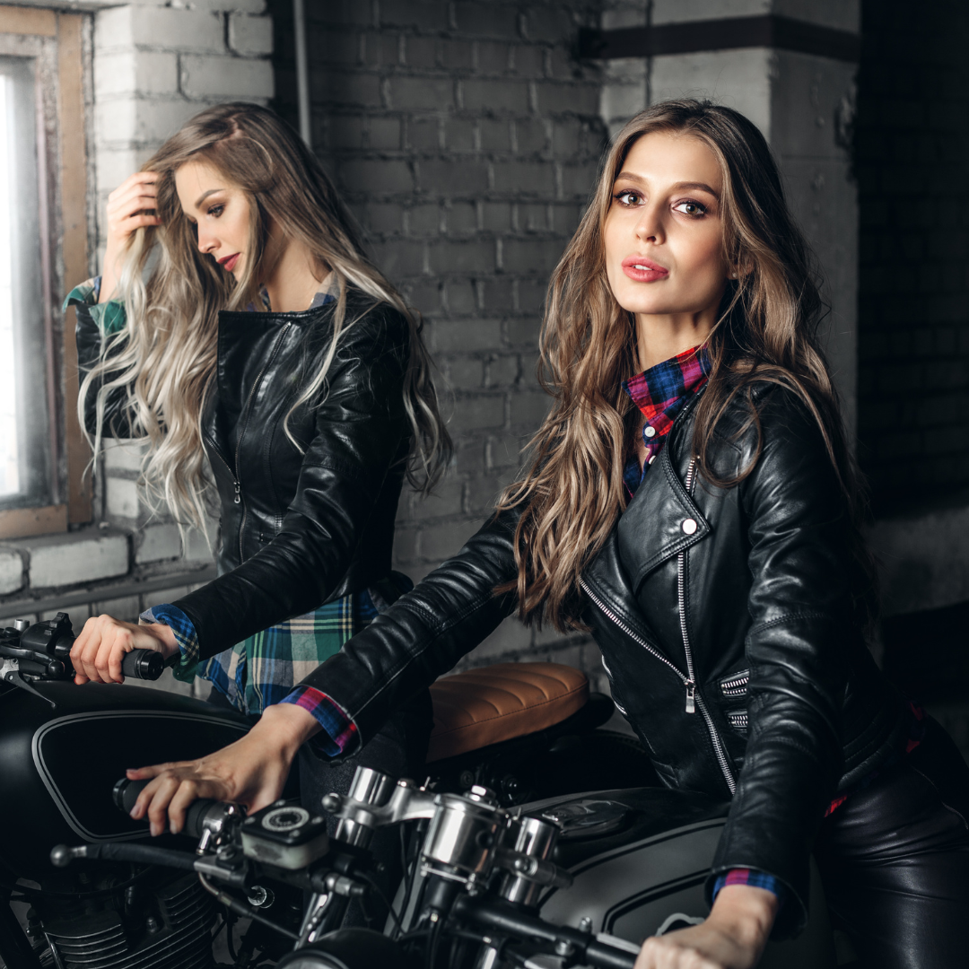 Two biker girls posing next to a motorcycle in leather jackets.